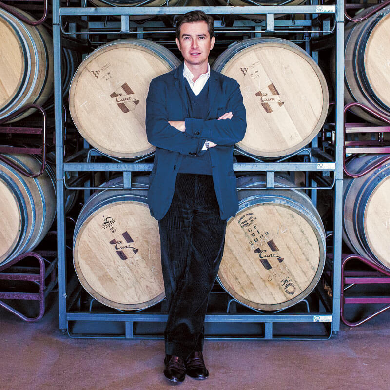 Victor Urrutia - CEO of CVNE dressed in a casual blue jacket standing in front of stacks of barrels ageing in the winery in Haro, Rioja