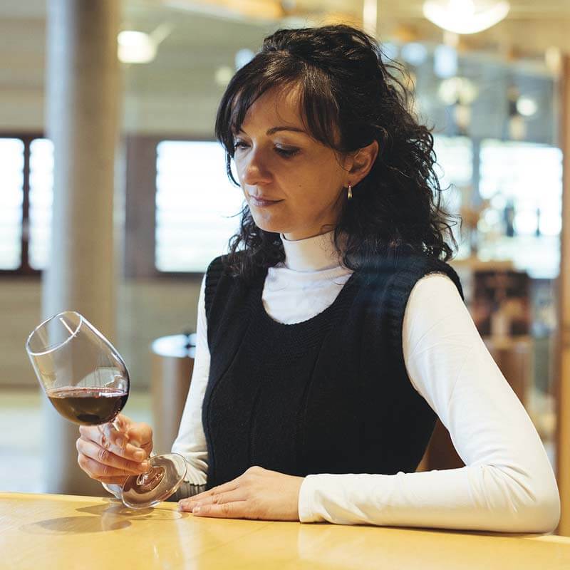 Eva de Benito, Head Winemaker of Viña Real in the Rioja Alavesa, stands wearing a black and white top, while examining the colour of a red wine in a glass tilted against a pale wooden counter top