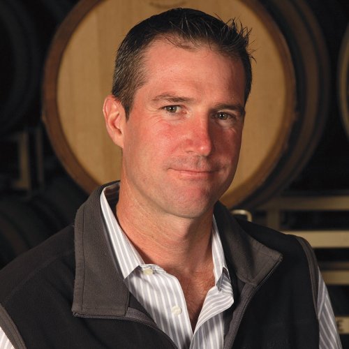 TJ Evans - Pinot Noir Winemaker - wearing a brown body warmer, stood inside the winery in front of stacked barrels
