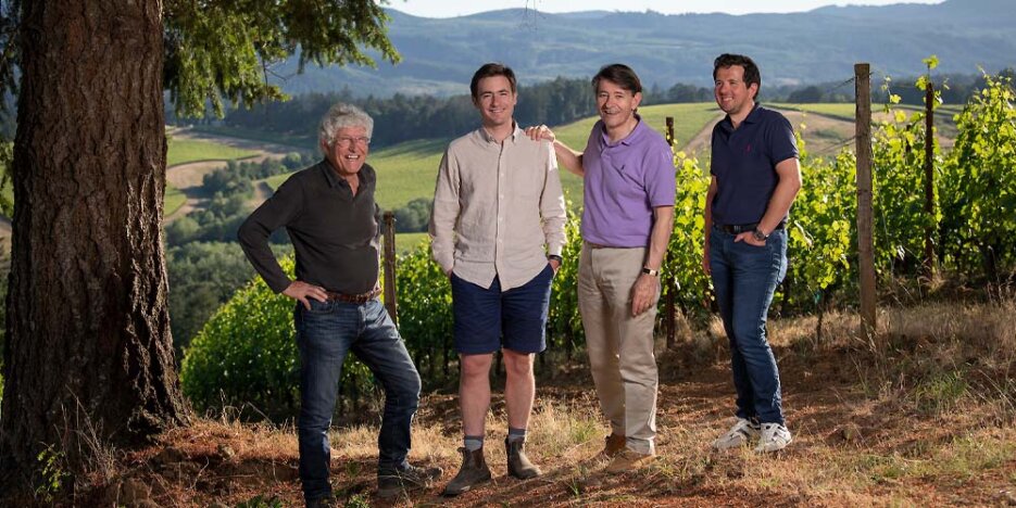 Four smiling men of different ages stand beneath a tree at the top of a vineyard which sweeps gently down the hill behind them. The man on the left standing with his hands on his hips has grey curly hair, a dark top and dark jeans. In the middle are son and father, the father in a purple polo shirt and beige chinos has his hand on his son's beige shirted shoulder. On the right end another dark haired man in a navy top and jeans, his hand in one pocket.