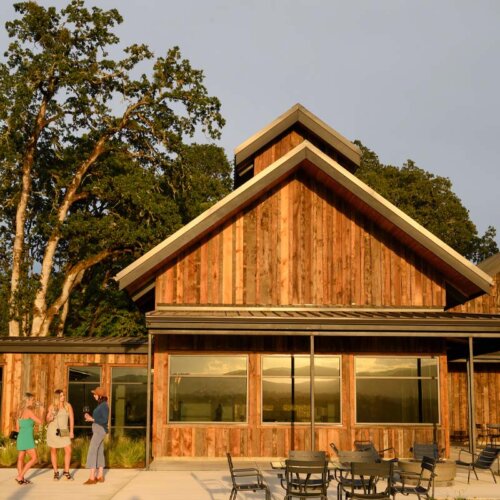 Three young ladies stand outside a red wood clad winery visitor centre, which has three simple large windows almost completely filling the ground floor visible under an overhang roof, above the wooden planked side of the building rises to an apex with a strong roofline, all topped by a smaller similar shaped apex structure in the middle of the roof ridge. Dark metal chairs and tables are in front of the building on the concrete patio.