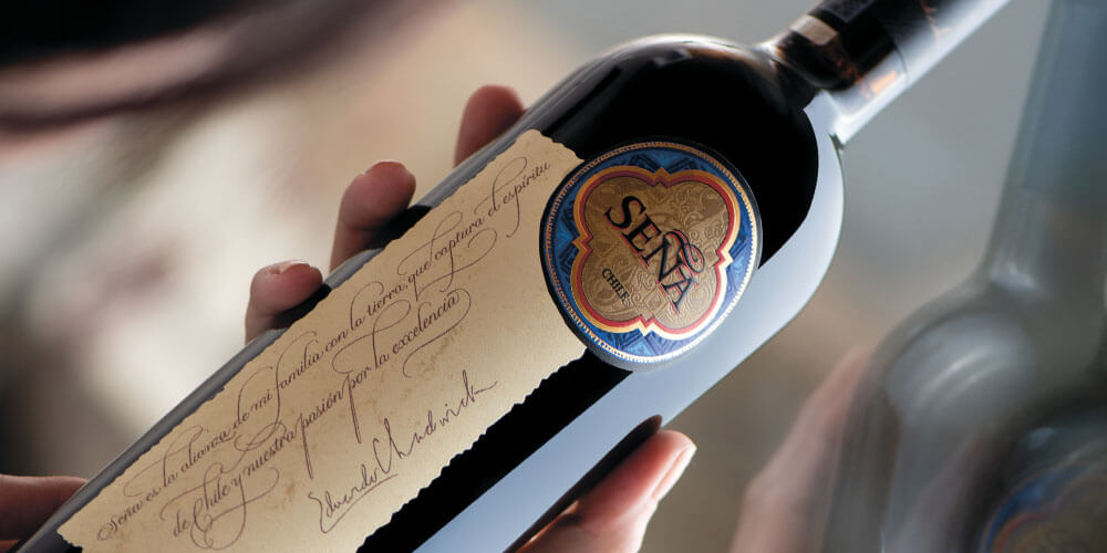 a lady's hand holds a dark glass bottle of Seña, showing clearly the cream coloured label with handwriting style spanish writing on, and a rich blue and gold round native south american style Seña design in the corner. The bottle is held against a glass window in which it is reflected.