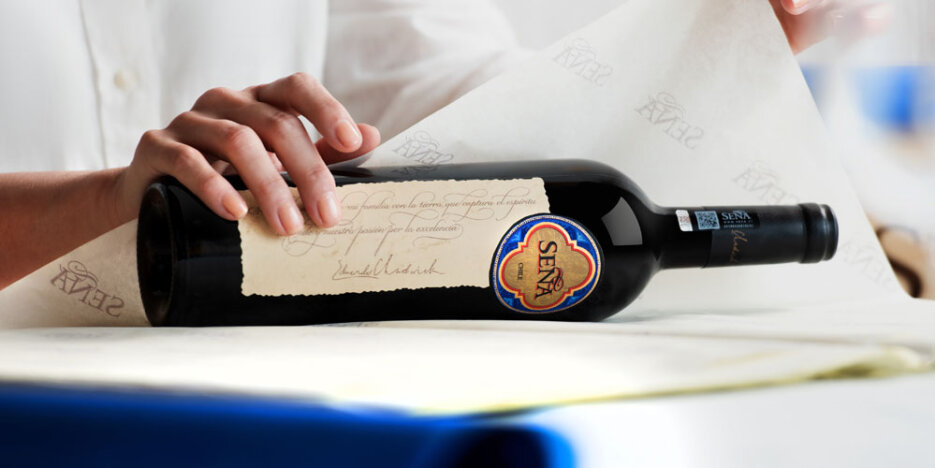 A bottle of Seña wine lying on its side, being carefully wrapped by hand in white tissue paper upon which are Seña logos.