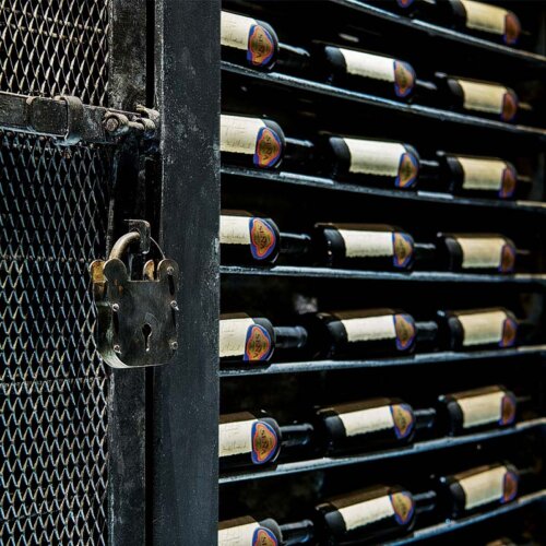 rows of bottles of Sena wine lying on side, one above the other in a metal vault on the wall, with the metal grid door pulled to one side, a sturdy padlock visible on the door.