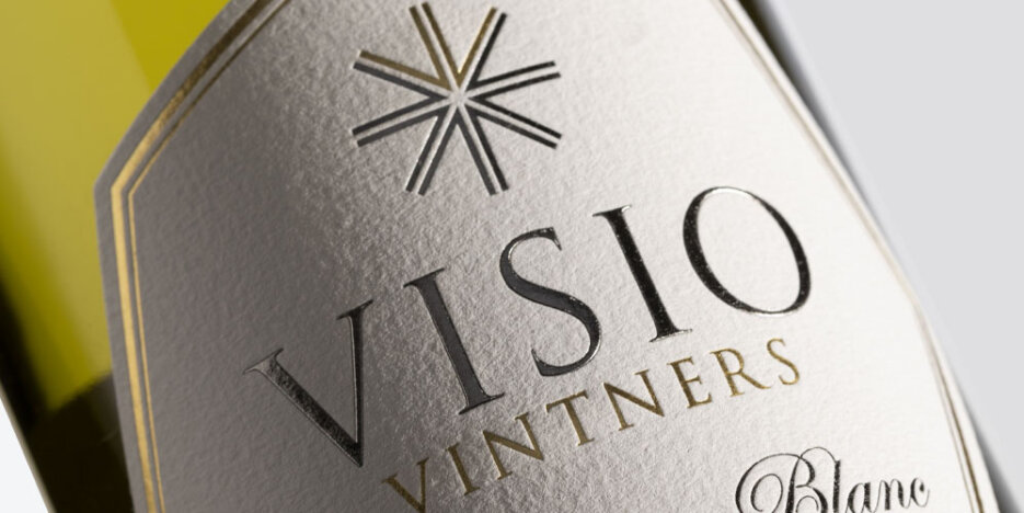 close of up a Visio Vintners wine label - the design has silver and gold metallic lettering on a textured white paper background