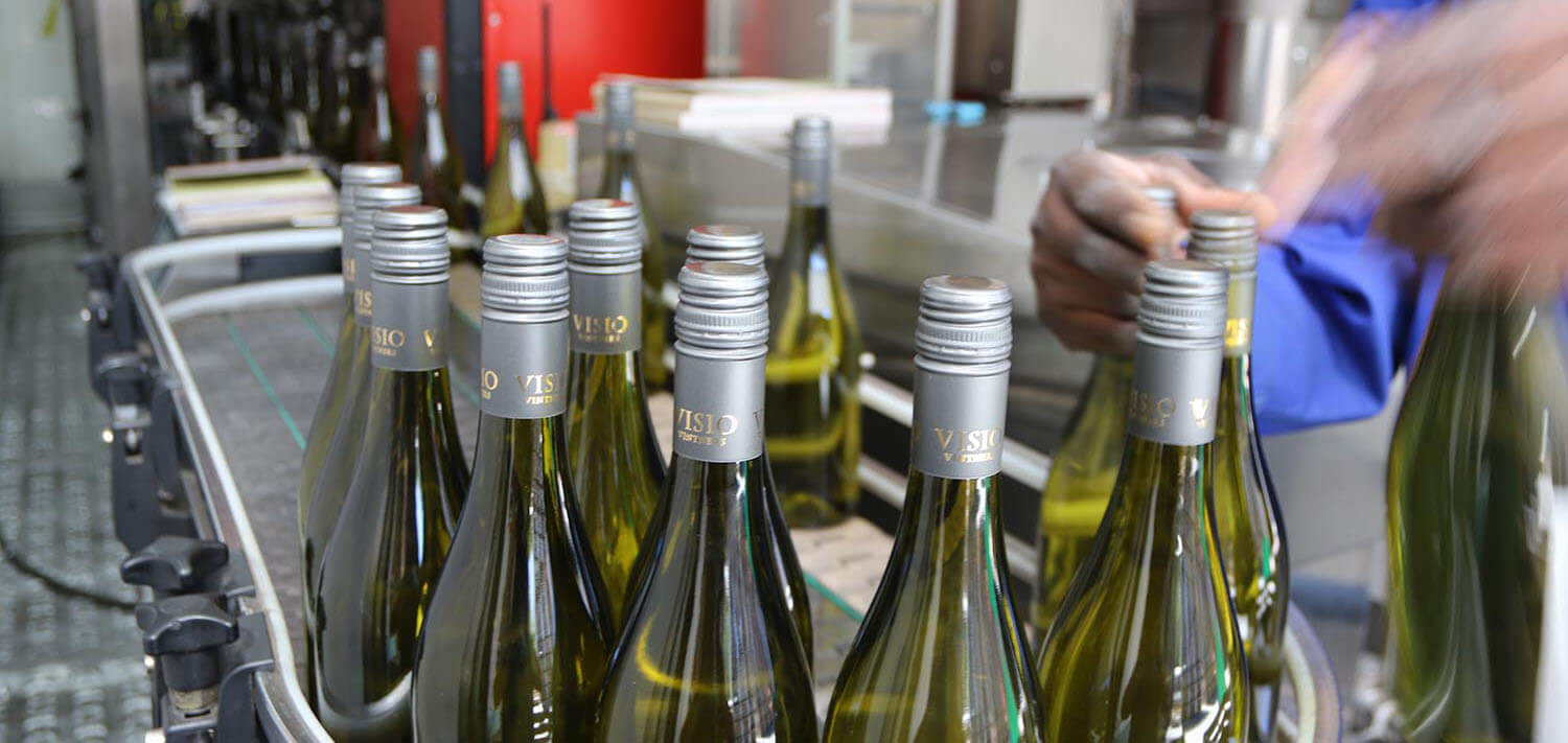 A line of green bottles with silver grey screw on bottle tops curling around a conveyor belt in the winery with a work on the right lifting bottles from the line