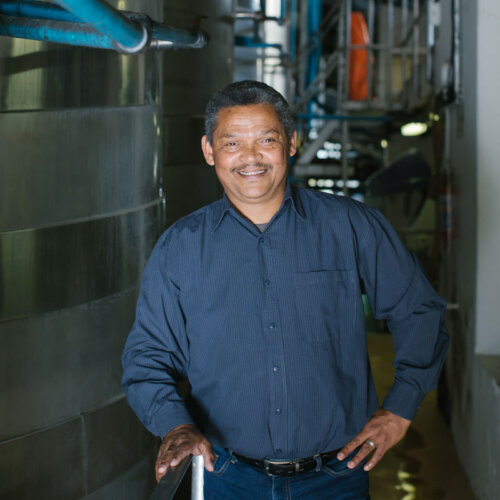black middle aged winery worker smiling at the camera, short cut dark grey hair, navy colour shirt and jeans, he's standing in the winery next to tall stainless steel fermenting tanks