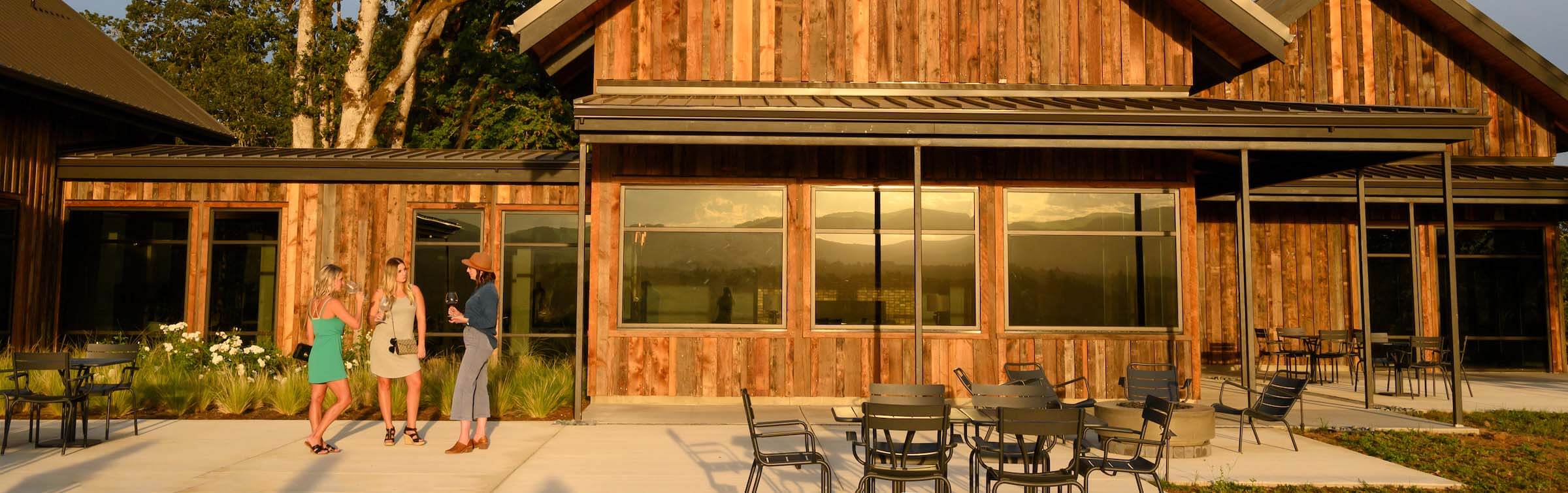 A close up of the ground floor of a red wood building with three large rectangular windows. In front are dark brown metal outdoor tables and chairs on a concrete floor, and three young ladies are stood on the left side of the image sipping wine and chatting.