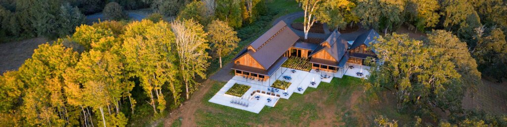 The winery visitor centre complex from the air above. A red wood walled set of angular buildings with dark roofs. The grey concrete outdoor seating areas form different shape rectangles in decreasing sizes, reflecting the different sizes of the building sections. Trees stand tall around the side and behind.