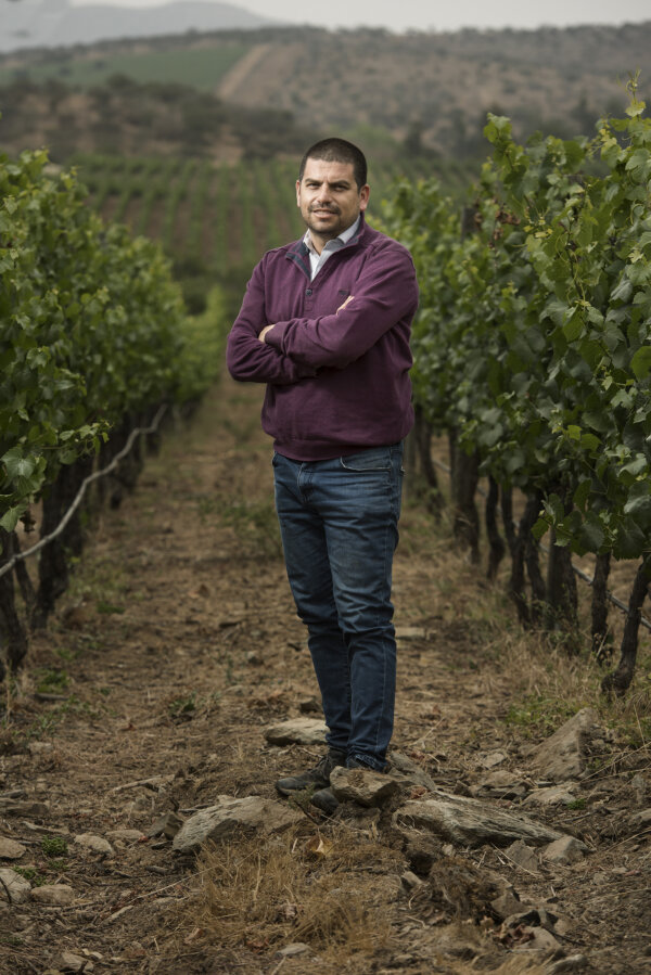 Carlos in a long sleeve plum red top and blue jeans, and short cropped dark hair, stands between a row of grape vines with his arms crossed