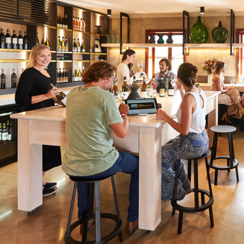 People sat on tall dark stools, around a large tall white table tasting wine. Shelves of wine to the side.