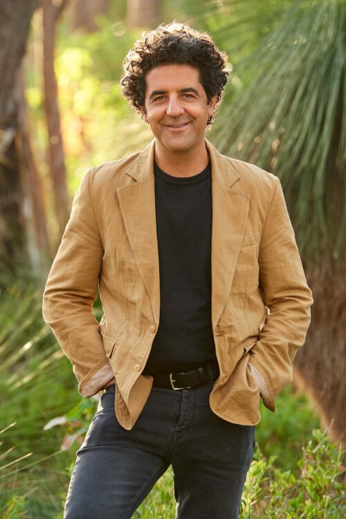 Larry Cherubino with a head of rich curly dark hair, smiles to the camera. He's wearing a tan jacket over a black round neck tshirt.
