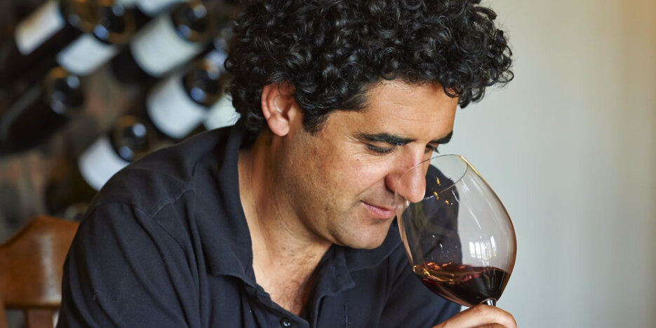 Larry Cherubino with a head of rich curly dark hair, wearing a black polo shirt, sniffing a large wine glass with red wine in