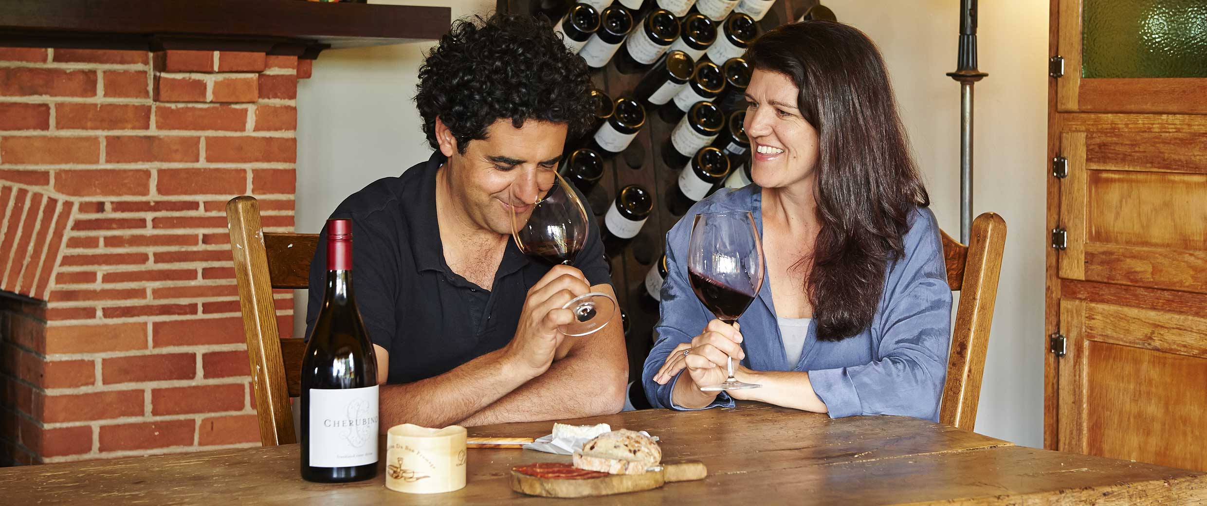 Larry with his dark curly hair, wearing a black polo neck and his wife Edwina with long dark hair, wearing a mid blue shirt, sit at a wooden table upon which is a block of cheese, a bottle of red wine and they are sampling. A red brick fireplace is behind on the left, and a large wine rack is behind them.
