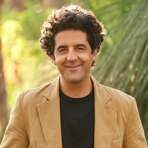 Larry Cherubino with a head of rich curly dark hair, smiles to the camera. He's wearing a tan jacket over a black round neck tshirt.